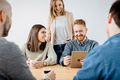Verschiedene Mitarbeitende sitzen und stehen um einen Tisch und schauen auf ein Tablet