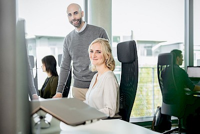 Zwei Mitarbeitende am Schreibtisch schauen in die Kamera