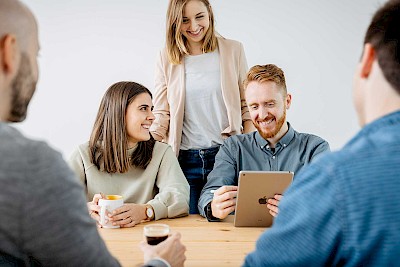 Drie werknemers kijken naar een tablet, zitten aan de vergadertafel, twee andere werknemers zitten ertegenover